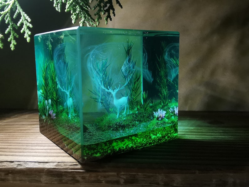 Puede incluir: Un cubo de resina transparente que muestra una escena de bosque en miniatura. Dos ciervos blancos se alzan entre plantas y flores verdes, con un efecto brumoso y et&eacute;reo. La obra de arte est&aacute; iluminada con un suave brillo verde.