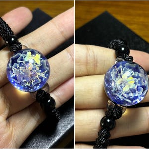 May include: A round, blue glass pendant with a floral design and a black cord. The pendant has a clear, bubble-like appearance with a detailed, colorful floral pattern inside. The cord is made of woven black material.