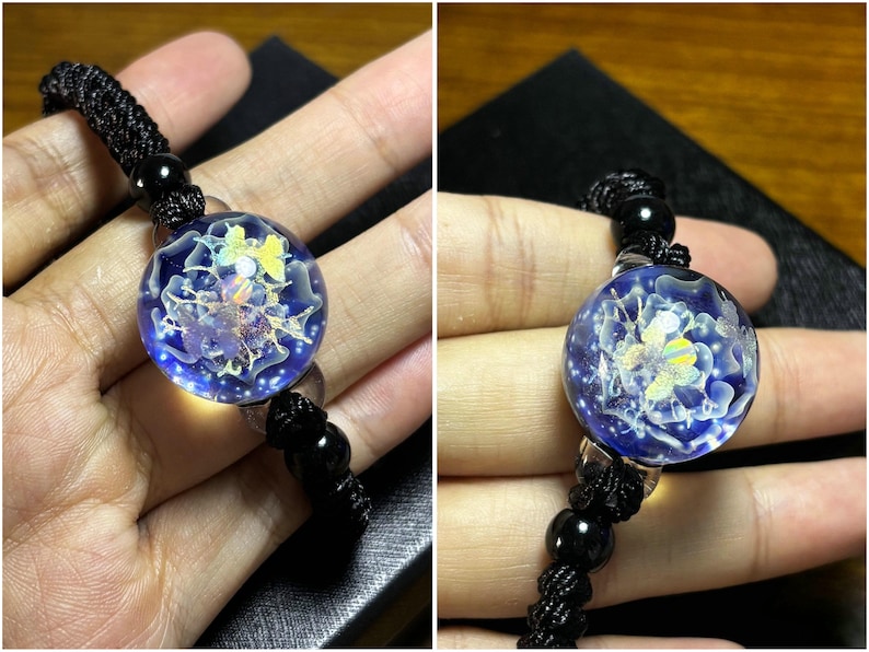 May include: A round, blue glass pendant with a floral design and a black cord. The pendant has a clear, bubble-like appearance with a detailed, colorful floral pattern inside. The cord is made of woven black material.