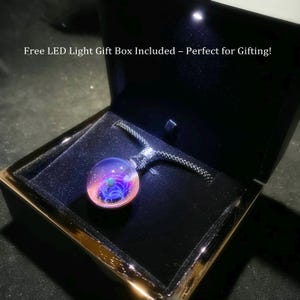 May include: A round, glass pendant necklace with a galaxy design, featuring shades of pink, purple, and blue. The pendant is displayed in an open, black gift box with a black cord. The box has a gold trim and a built-in LED light.