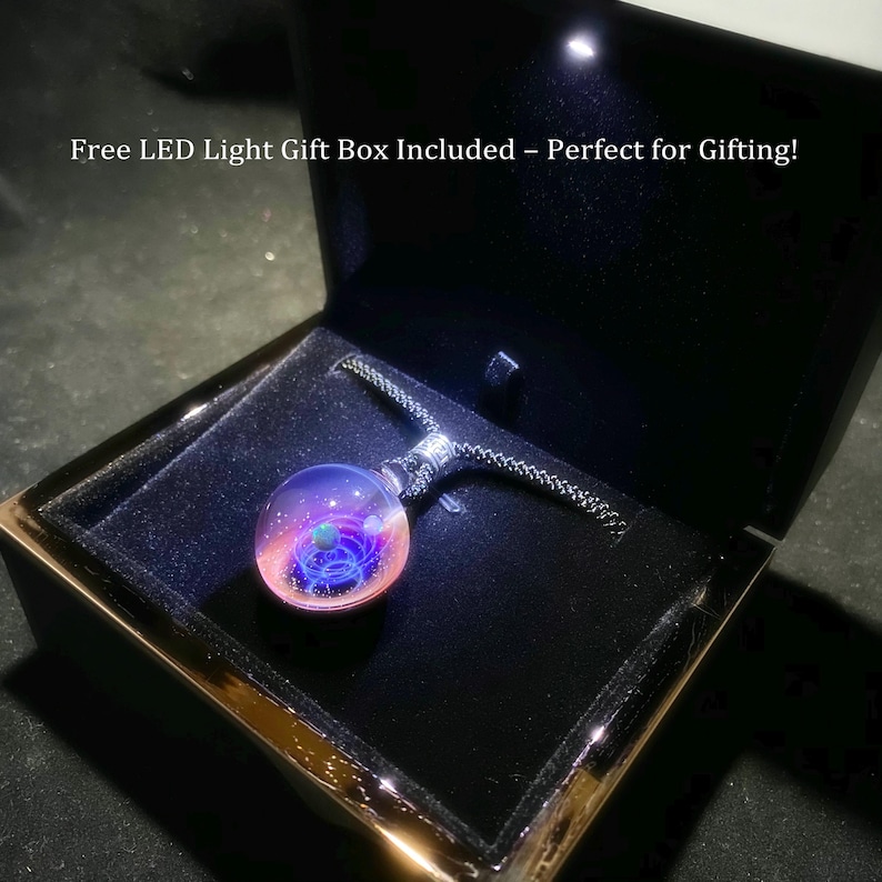 May include: A round, glass pendant necklace with a galaxy design, featuring shades of pink, purple, and blue. The pendant is displayed in an open, black gift box with a black cord. The box has a gold trim and a built-in LED light.