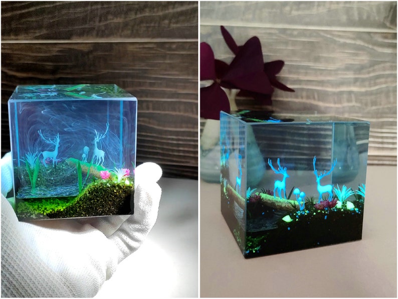 Puede incluir: Un cubo de resina transparente que presenta una escena de bosque en miniatura con dos ciervos, plantas y hongos. La escena est&aacute; iluminada con un brillo azul, creando un efecto m&aacute;gico. El cubo es sostenido por una mano enguantada.