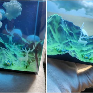 Puede incluir: Diorama de paisaje en miniatura con un ciervo, monta&ntilde;as y nubes. Las monta&ntilde;as son de un verde vibrante y el cielo es azul profundo. El ciervo es blanco y las nubes son esponjosas. La escena est&aacute; encerrada en resina transparente.