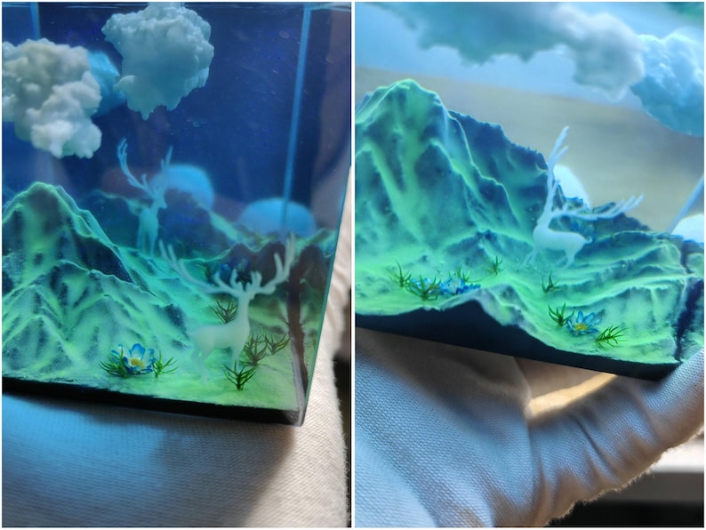Puede incluir: Diorama de paisaje en miniatura con un ciervo, monta&ntilde;as y nubes. Las monta&ntilde;as son de un verde vibrante y el cielo es azul profundo. El ciervo es blanco y las nubes son esponjosas. La escena est&aacute; encerrada en resina transparente.