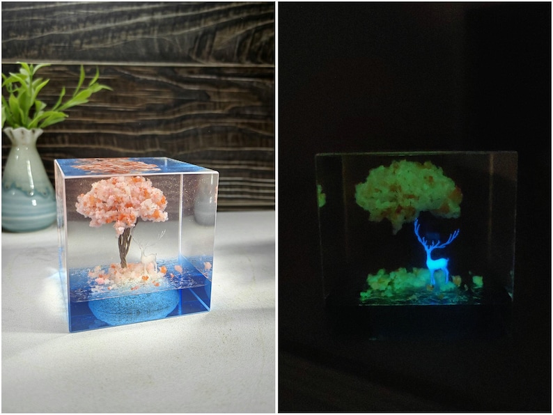 Puede incluir: Un cubo de resina transparente con un &aacute;rbol en miniatura de flores de cerezo rosas y una base azul. El &aacute;rbol est&aacute; dentro de un bloque de resina transparente. El cubo brilla en la oscuridad, revelando un ciervo.