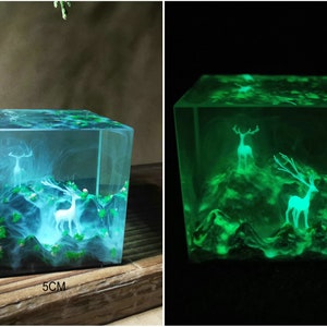Puede incluir: Un cubo de resina transparente, de 5 cm por lado, encapsula un paisaje en miniatura con figuras de ciervos blancos y follaje verde. La escena brilla con una suave luz azul, creando un efecto m&iacute;stico. El cubo est&aacute; sobre una superficie de madera.