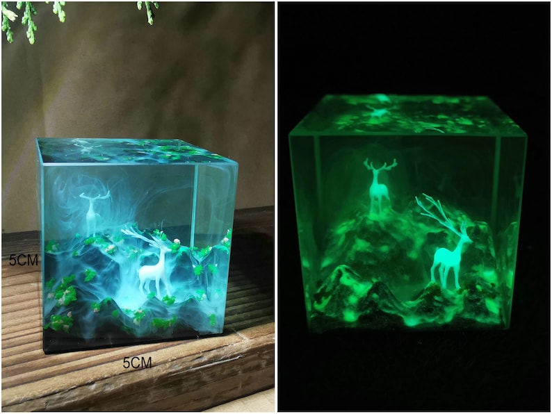 Puede incluir: Un cubo de resina transparente, de 5 cm por lado, encapsula un paisaje en miniatura con figuras de ciervos blancos y follaje verde. La escena brilla con una suave luz azul, creando un efecto m&iacute;stico. El cubo est&aacute; sobre una superficie de madera.