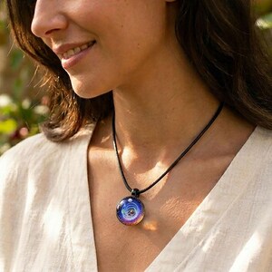 May include: A round pendant necklace with a black cord. The pendant features a colorful, swirling design with shades of blue, purple, and white. The person in the image is wearing a cream-colored top.
