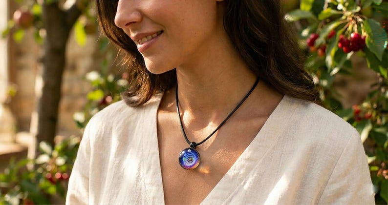 May include: A round pendant necklace with a black cord. The pendant features a colorful, swirling design with shades of blue, purple, and white. The person in the image is wearing a cream-colored top.