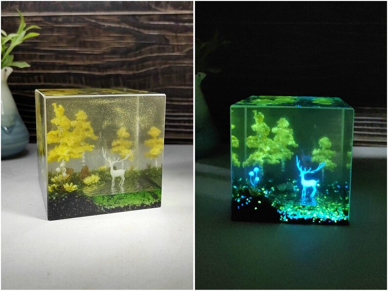 Puede incluir: Un cubo de resina transparente con una escena de bosque en miniatura. La escena incluye &aacute;rboles amarillos, un ciervo blanco y una base verde. El cubo brilla en verde en la oscuridad, con el ciervo y los &aacute;rboles iluminados.