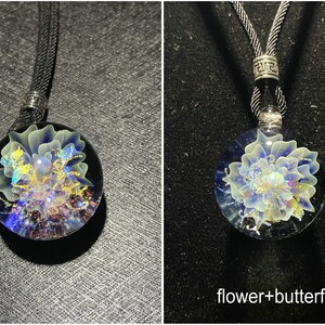 May include: A round glass pendant necklace with a flower and butterfly design. The pendant features a blue and white flower with iridescent accents, suspended within a clear glass orb. The necklace has a black cord.