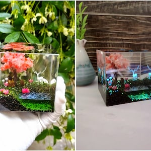 Puede incluir: Un cubo de resina transparente que presenta un paisaje en miniatura con un &aacute;rbol rosa, ciervos blancos y un peque&ntilde;o estanque. La escena incluye musgo verde, flores rosas y hongos azules brillantes, creando un diorama caprichoso e iluminado.