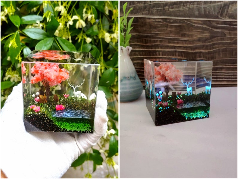 Puede incluir: Un cubo de resina transparente que presenta un paisaje en miniatura con un &aacute;rbol rosa, ciervos blancos y un peque&ntilde;o estanque. La escena incluye musgo verde, flores rosas y hongos azules brillantes, creando un diorama caprichoso e iluminado.