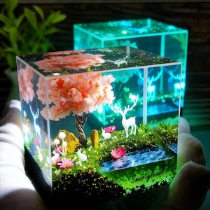 Puede incluir: Un cubo de resina transparente con una escena de paisaje en miniatura. Presenta un &aacute;rbol de flores rosas, figuras de ciervos blancos y un peque&ntilde;o arroyo. El texto "Smartylecoul" es visible.
