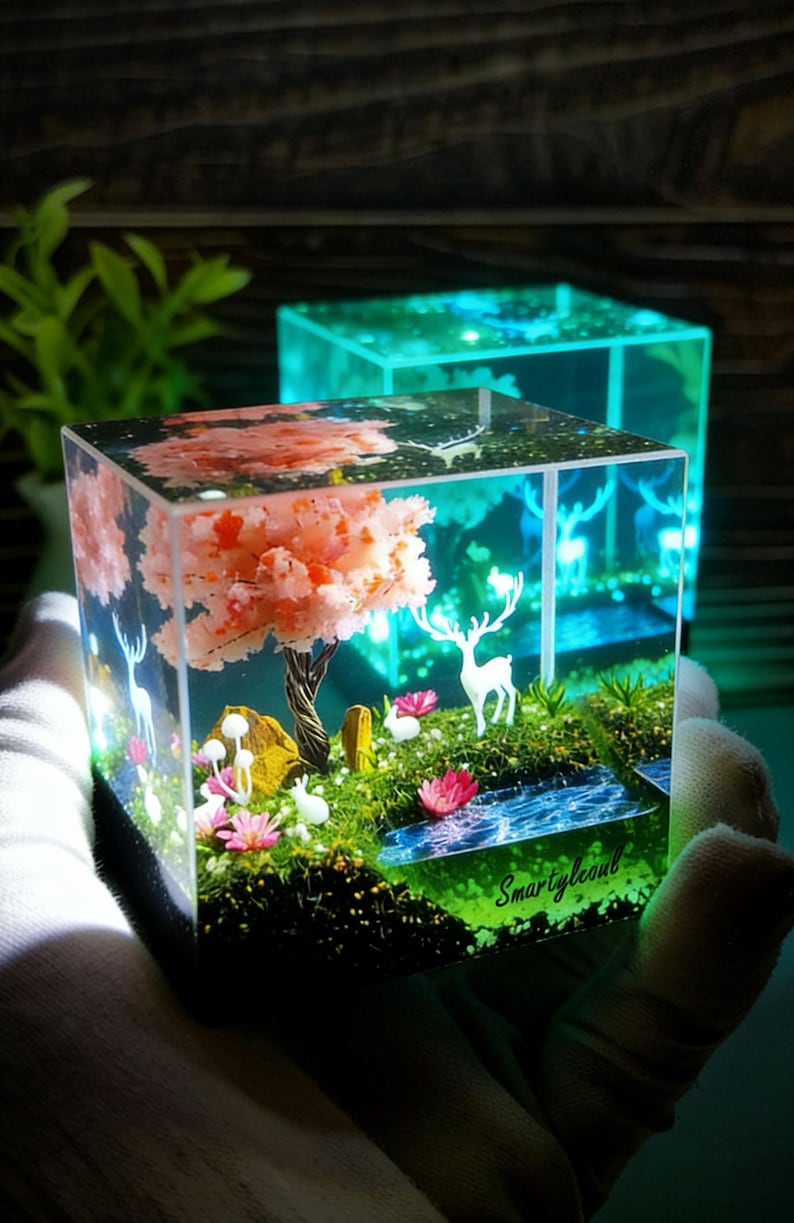 Puede incluir: Un cubo de resina transparente con una escena de paisaje en miniatura. Presenta un &aacute;rbol de flores rosas, figuras de ciervos blancos y un peque&ntilde;o arroyo. El texto "Smartylecoul" es visible.