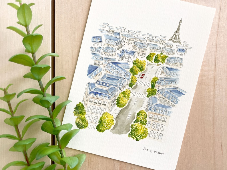 K&ouml;nnte beinhalten: Ein Aquarellbild einer Stra&szlig;enszene in Paris, Frankreich. Das Bild zeigt eine Stra&szlig;e, die von Geb&auml;uden, B&auml;umen und einem roten Auto ges&auml;umt ist. Der Eiffelturm ist in der Ferne sichtbar. Der Text "Paris, France" steht am unteren Rand des Bildes.