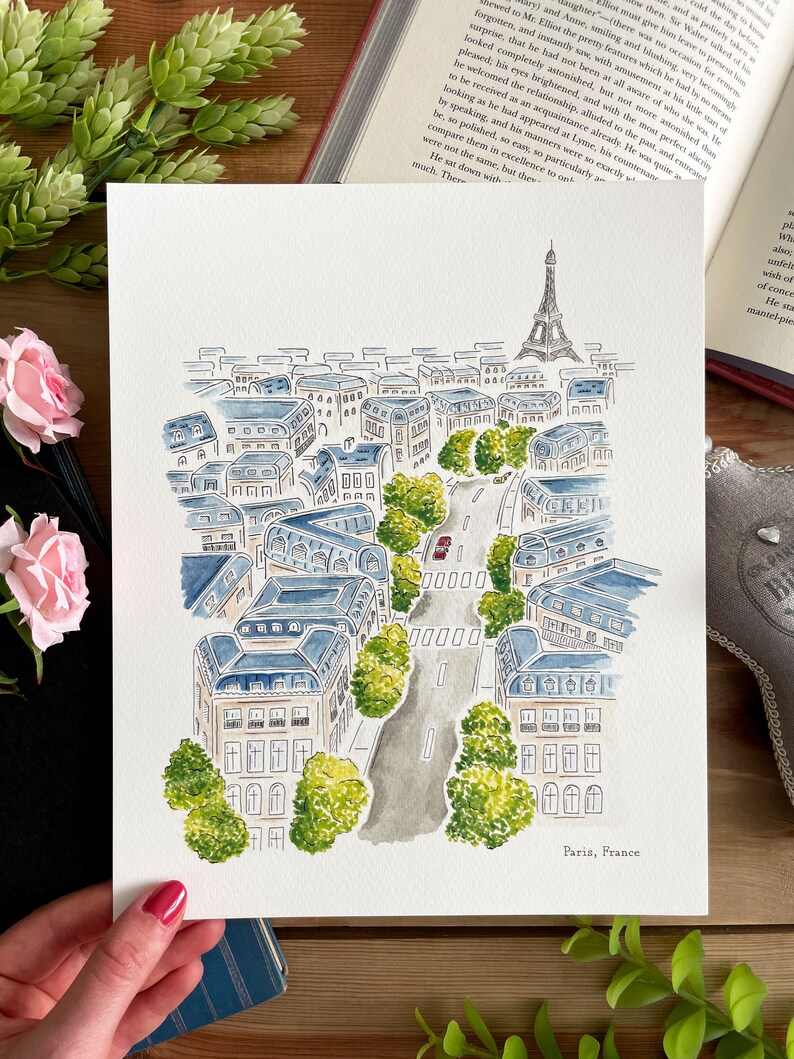 Puede incluir: Pintura a la acuarela de Par&iacute;s, Francia, con la Torre Eiffel y edificios de la ciudad con tejados azules. Una carretera con un coche rojo serpentea por la ciudad, bordeada de &aacute;rboles verdes. El texto "Paris, France" est&aacute; en la parte inferior.