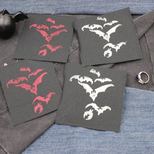 May include: Four square black fabric patches featuring a screen-printed design of a flock of bats in flight. Two patches display the bat motif in a deep red ink, while the other two patches feature the same bat design in a bright white ink.