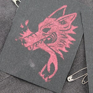 May include: A rectangular black fabric patch featuring a screen-printed graphic of a stylized wolf head in a muted red color. The patch has a textured weave and is attached to a dark denim surface with a silver-toned metal safety pin.