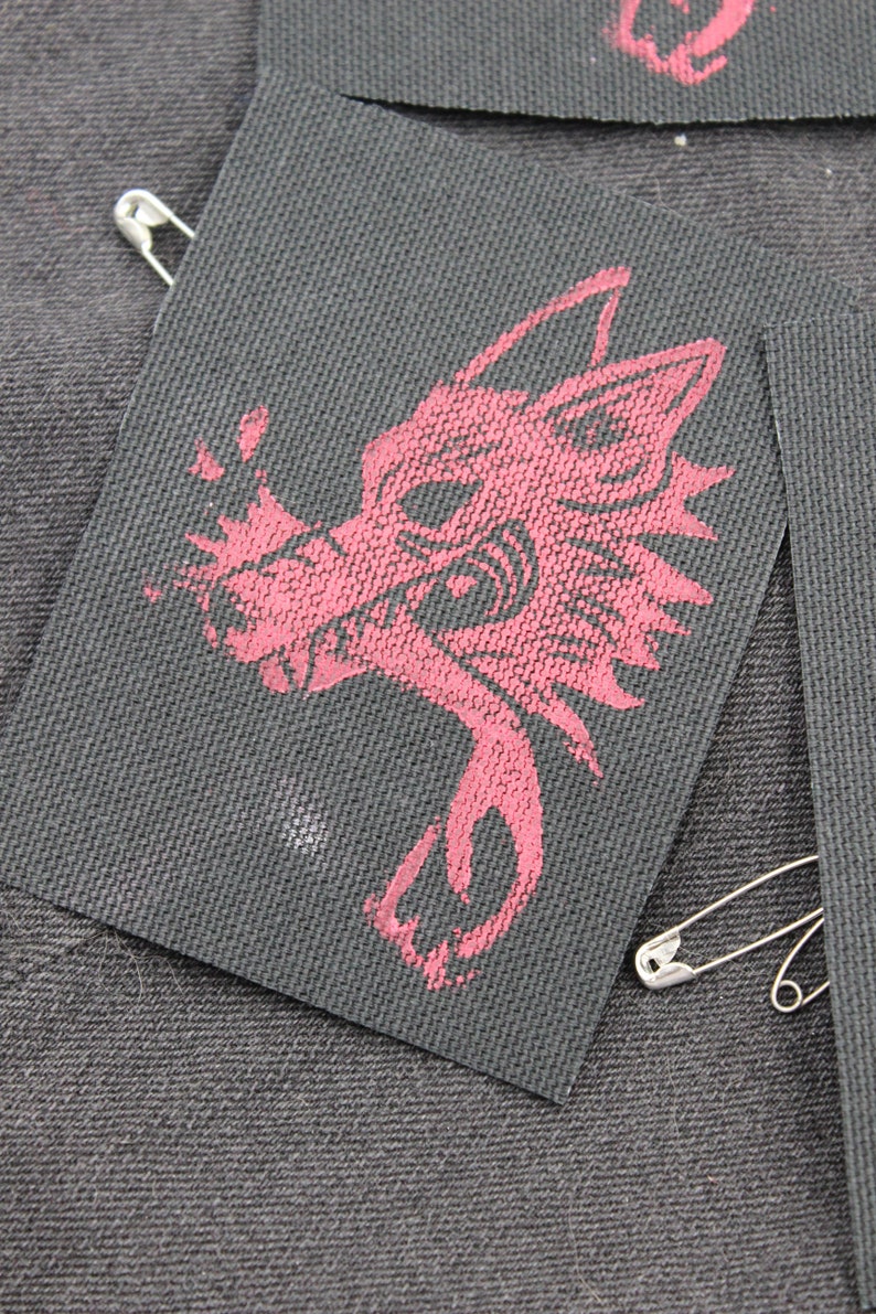 May include: A rectangular black fabric patch featuring a screen-printed graphic of a stylized wolf head in a muted red color. The patch has a textured weave and is attached to a dark denim surface with a silver-toned metal safety pin.