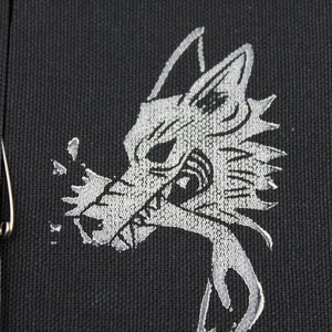 May include: A black fabric patch featuring a silver embroidered graphic of a stylized wolf head with sharp features and a jagged mane. The wolf is depicted with its mouth open, showing teeth, and a silver metallic safety pin is attached to the side.