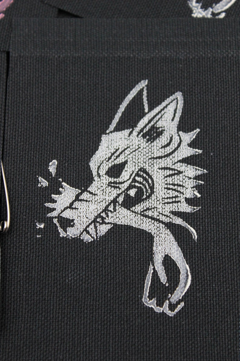 May include: A black fabric patch featuring a silver embroidered graphic of a stylized wolf head with sharp features and a jagged mane. The wolf is depicted with its mouth open, showing teeth, and a silver metallic safety pin is attached to the side.
