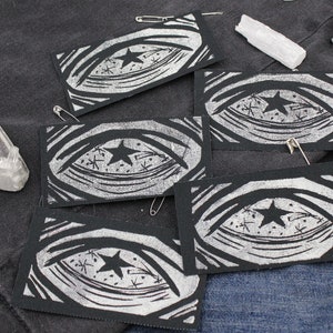May include: Four rectangular black fabric patches featuring a white screen-printed graphic of an eye with a star in the center and radiating lines. The patches are arranged on a dark fabric surface alongside two clear crystals and metal safety pins.