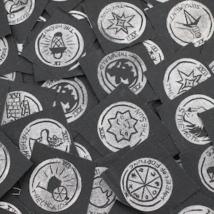 May include: A collection of square black fabric patches featuring white screen-printed tarot card designs. Each patch displays a unique circular illustration with text such as The Sun, The Hermit, The Chariot, The World, and Strength, arranged in a scattered overlapping pile.