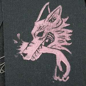 May include: A rectangular black fabric patch featuring a screen-printed graphic of a stylized wolf head in a light pink color. The wolf design shows sharp features and a jagged mane, with the ink texture visible against the woven fabric surface.