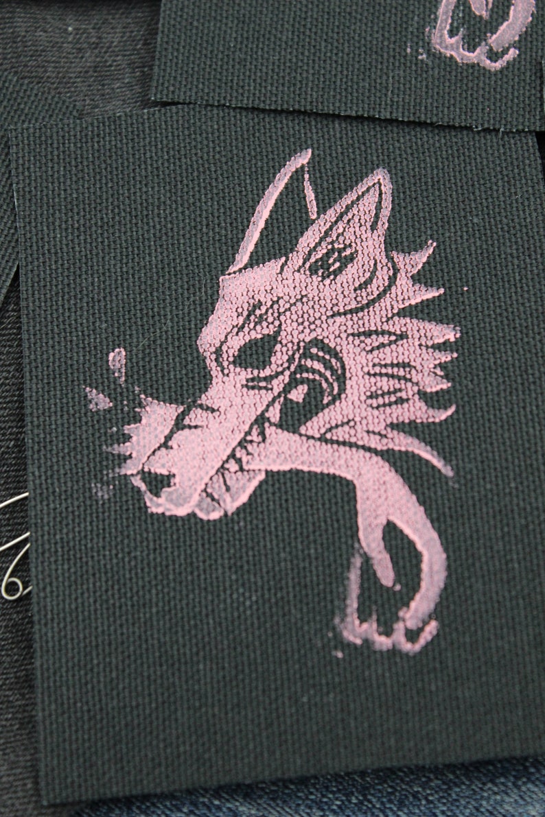 May include: A rectangular black fabric patch featuring a screen-printed graphic of a stylized wolf head in a light pink color. The wolf design shows sharp features and a jagged mane, with the ink texture visible against the woven fabric surface.