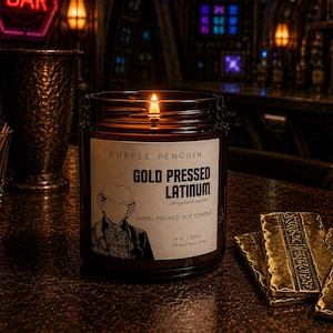 Peut inclure: Une bougie en pot de verre brun avec une mèche allumée et une étiquette blanche indiquant Purple Penguin Gold Pressed Latinum, Cardamom-Vetiver, Hand Poured Soy Candle, 9 oz, 255 g, 50 hour burn time, avec une illustration au trait d'un humanoïde.