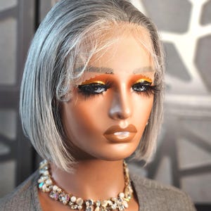 May include: A mannequin head with a short, gray wig and a sparkling necklace. The wig has a natural-looking hairline and is styled in a bob. The mannequin has dramatic eye makeup with long lashes and gold eyeshadow. The necklace features various clear and iridescent stones.
