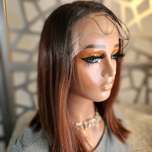 May include: A mannequin head displaying a brown and auburn ombre wig with a lace front. The wig is styled in a shoulder-length bob. The mannequin is wearing a gray shirt and a gold necklace.