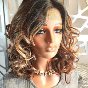 May include: A mannequin head displaying a brown and blonde curly wig. The wig has dark roots and lighter highlights. The mannequin is wearing a gray ribbed shirt and a decorative necklace. The background is a neutral color.
