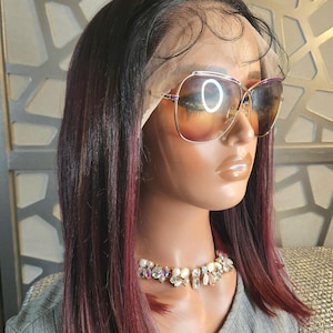 May include: A mannequin head with a burgundy and black wig, wearing oversized sunglasses with a gold frame and a jeweled necklace. The wig has a blunt cut and is styled straight. The background has a geometric pattern.