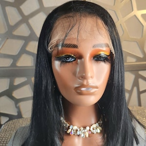 May include: A mannequin head with a black wig, styled in a shoulder-length cut. The mannequin has dramatic eye makeup with orange and yellow eyeshadow and a jeweled necklace. The mannequin is wearing a gray top.