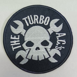 Puede incluir: Un parche bordado en blanco y negro con un diseño de calavera y huesos cruzados. El texto "THE TURBO A.C.'s" está escrito alrededor de la calavera.