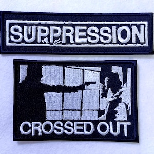 May include: Two black embroidered patches with white stitching. The top patch says "SUPPRESSION" in all caps. The bottom patch has a graphic of a window with bars crossed out and says "CROSSED OUT" in all caps.