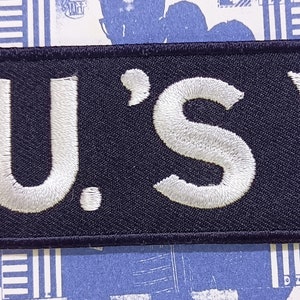 May include: A black embroidered patch with white lettering that reads "F.U.'S" and a white triangle.