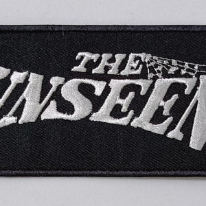 Peut inclure: Un patch en tissu noir avec du texte brodé blanc qui dit "THE UNSEEN".