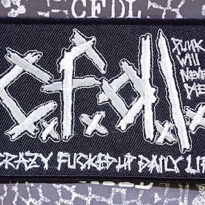 Może przedstawiać: Czarna prostokątna naszywka z białym haftowanym tekstem i grafiką. Na naszywce widnieje napis "CFDL" w stylizowanej czcionce, z frazą "CRAZY FUCKED UP DAILY LIFE" poniżej. Tekst "PUNK WILL NEVER DIE!" jest również zawarty.