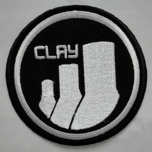 以下が含まれることがあります： 「CLAY」という文字と、白い靴下が3つ円形に並んだ、黒と白の刺繍パッチです。