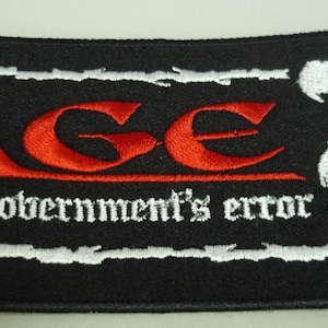 以下が含まれることがあります： 黒と白の刺繍パッチで、赤い「AGE」ロゴと「armed government's error」のテキストが書かれています。パッチの右側には、白い鳥のような生き物がいます。
