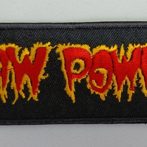 Może przedstawiać: Czarna prostokątna naszywka z wyhaftowanym napisem "RAW POWER" w kolorze czerwonym z żółtymi płomieniami. Naszywka jest przeznaczona do naszywania na odzież lub torby, z odważną, surową czcionką.