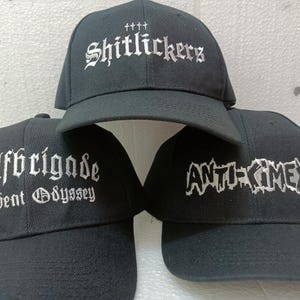 以下が含まれることがあります： 白い刺繍の文字が入った黒い野球帽が3つ。帽子には「Shitlickers」、「Wolfbrigade A-Bent Odyssey」、「Anti-Cimex」の文字が書かれている。