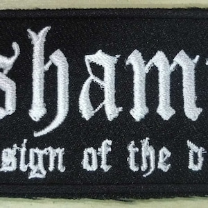 Puede incluir: Parche rectangular negro con texto bordado en blanco. La palabra "Dishammer" está en una fuente gótica sobre la frase "under the sign of the d-beat mark". El parche es probablemente para una chaqueta o bolso.
