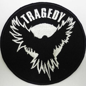 Op de afbeelding: Zwart-witte geborduurde patch met een gestileerd vogelontwerp en het woord "TRAGEDY" in witte letters.