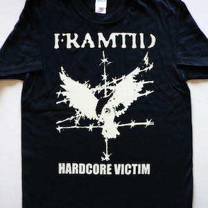 Peut inclure: T-shirt noir avec un motif blanc d'un oiseau aux ailes déployées, entouré de barbelés et d'étoiles. Le texte "FRAMTID" et "HARDCORE VICTIM" est imprimé en blanc.