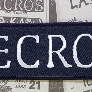 May include: A rectangular embroidered patch with a black background and a white stitched border. The word NECROS is embroidered in large, bold, white capital letters across the center of the patch, featuring a clean and simple sans-serif font style.