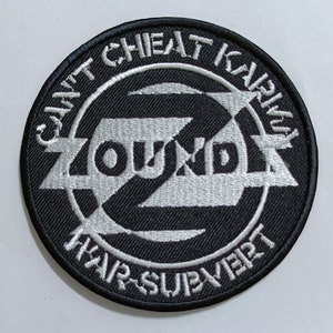 May include: A black and white embroidered patch with the text "ZOUND!" in a circle. The text "Can't Cheat Karma" is above the circle and "Fair Subvert" is below the circle.